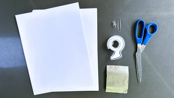 Zwei weiße Kartonblätter, eine Stecknadel, eine Büroklammer, Klebeband, ein kleiner Beutel und eine blaue Schere auf dunklem Untergrund.