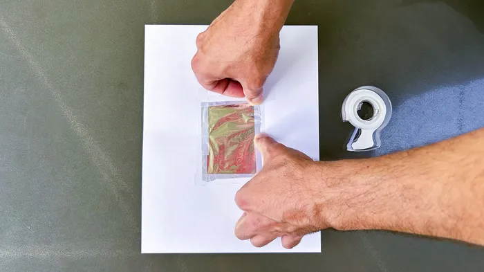 Hände befestigen ein kleines, farbiges Objekt mit transparentem Klebeband auf einem weißen Karton.