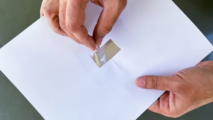 Hände platzieren eine glänzende Folie über einem rechteckigen Ausschnitt in einem weißen Karton.