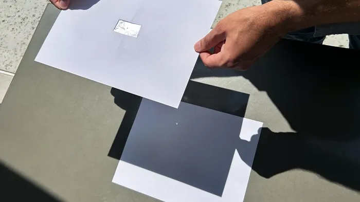 Zwei weiße Kartonblätter für einen Pinhole-Projektor, eines mit einer rechteckigen Öffnung und eines mit einem kleinen Loch.