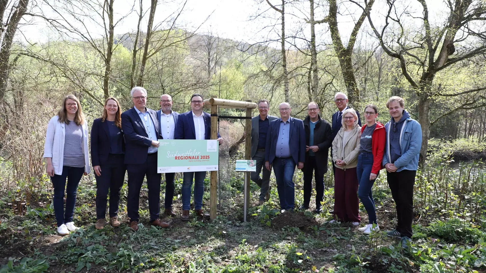 Eine Gruppe von zehn Personen steht vor einem Holzschild mit dem Aufdruck „Südwestfalen REGIONALE 2025“ in einer Waldlandschaft.