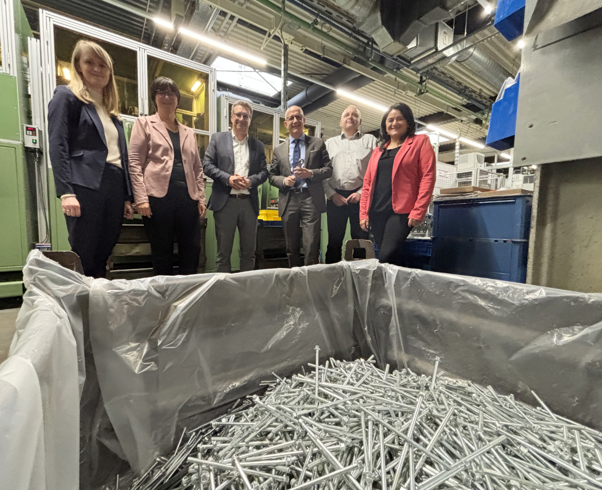 Sechs Personen stehen vor einem großen Behälter mit Metallteilen in einer Produktionshalle. Von links sind zu sehen: Johanna Muhl, Kirsten Kling, Stephan Schwarz, Ralf Schwarzkopf, Burkard Götz und Fatma Mendoza.