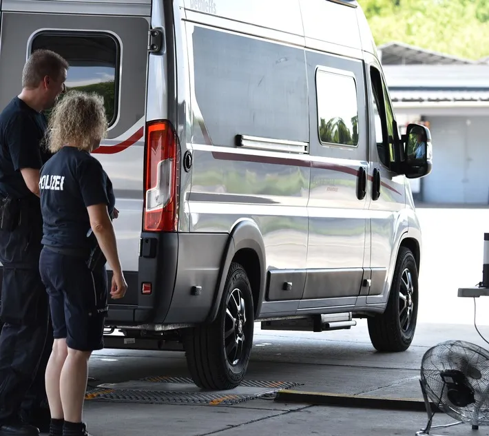 Polizeibeamte stehen neben einem grauen Wohnmobil, während dieses auf einer Waage steht. Im Hintergrund ist ein Ventilator und ein Gebäude zu sehen.