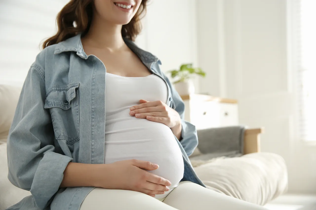 Eine lächelnde schwangere Frau sitzt auf einer Couch und hält sanft ihren Bauch.