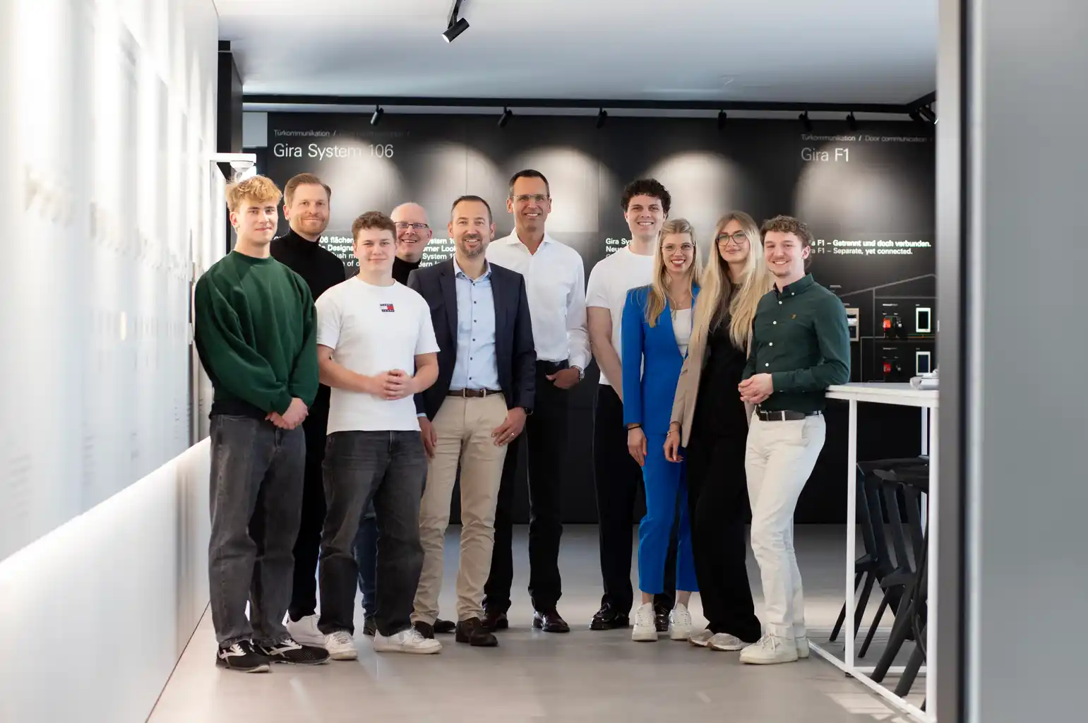 Gruppe von zehn jungen Berufstätigen und vier Führungskräften posiert in einem hellen Büroraum mit moderner Inneneinrichtung. Im Hintergrund sind Schilder mit der Aufschrift „Gira System 106“ und „Gira F1“ zu sehen.