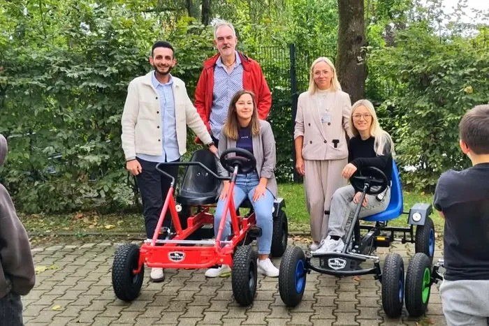 Fünf Personen posieren bei einer Spendenübergabe mit zwei kleinen Pedalkarts im Freien.