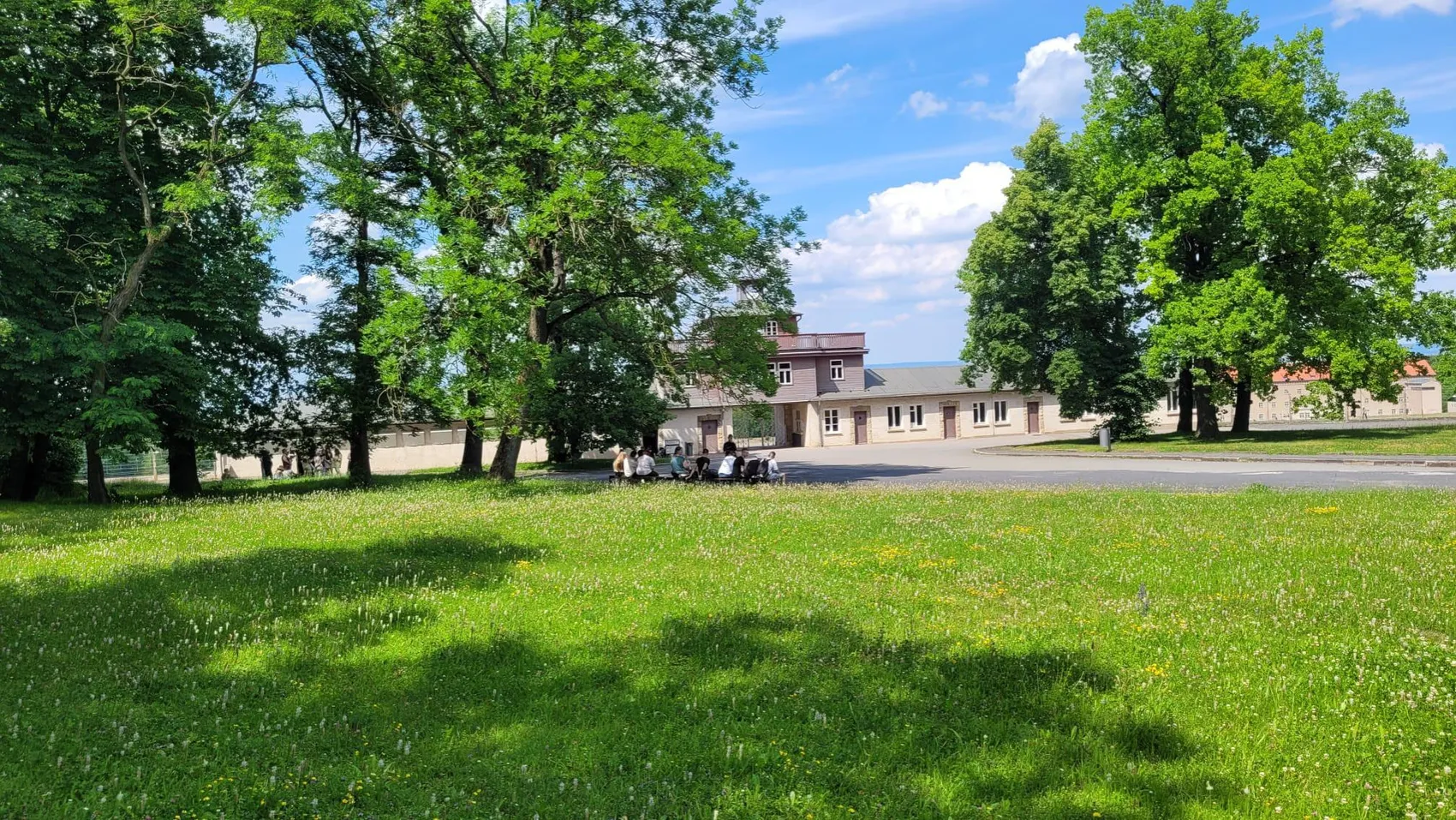 Ein helles Gebäude mit rotem Dach ist von Bäumen und einer grünen Wiese umgeben. Im Vordergrund sitzen Schüler auf einer Bank.