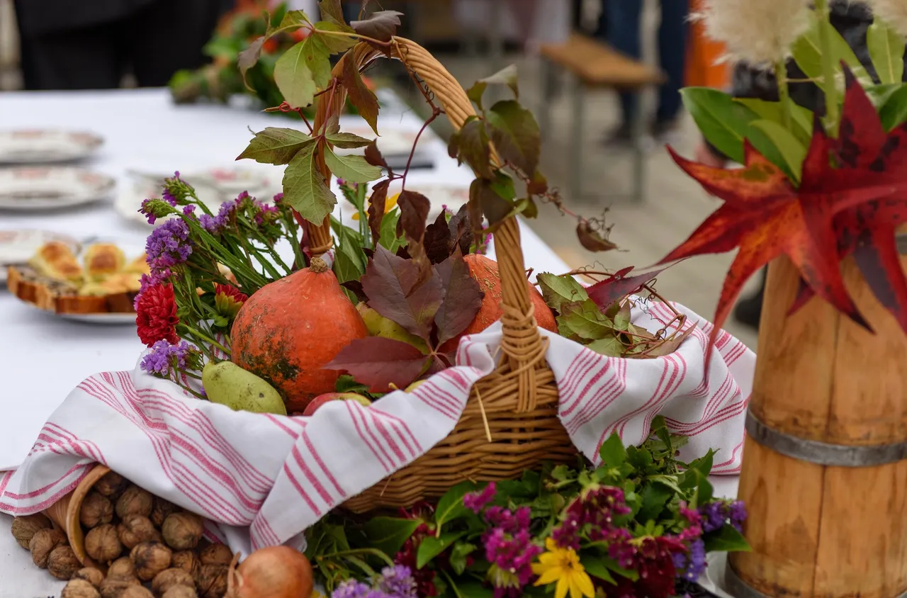 Ein geflochtener Korb mit einem Kürbis, Weintrauben und bunten Blumen auf einem rot-weiß gestreiften Tuch.