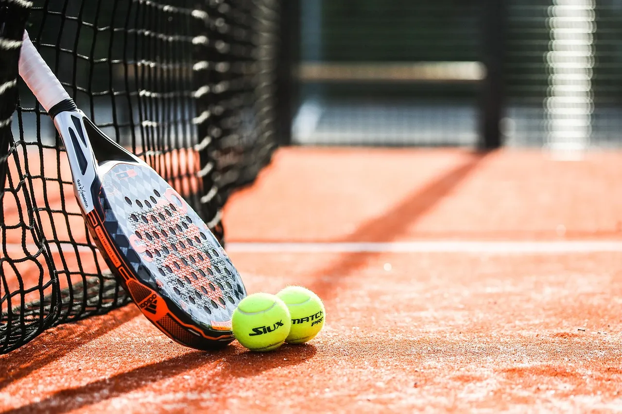 Ein Padel-Schläger lehnt an einem schwarzen Netz auf einem roten Sandplatz neben zwei gelben Bällen.