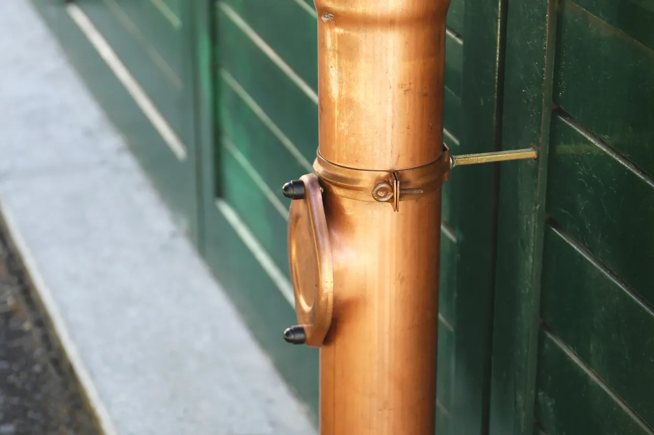 Kupfernes Fallrohr mit einer Revisionsöffnung an einer dunkelgrünen Wand.