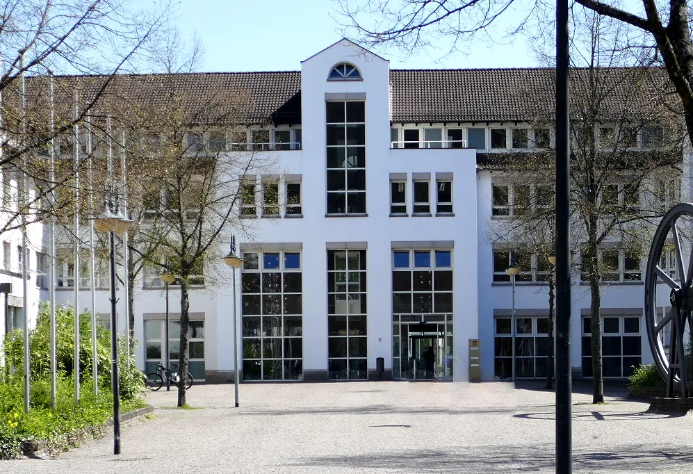 Weißer Verwaltungsbau mit großflächigen Fenstern und einem markanten Giebel, davor ein gepflasterter Platz mit Bäumen.