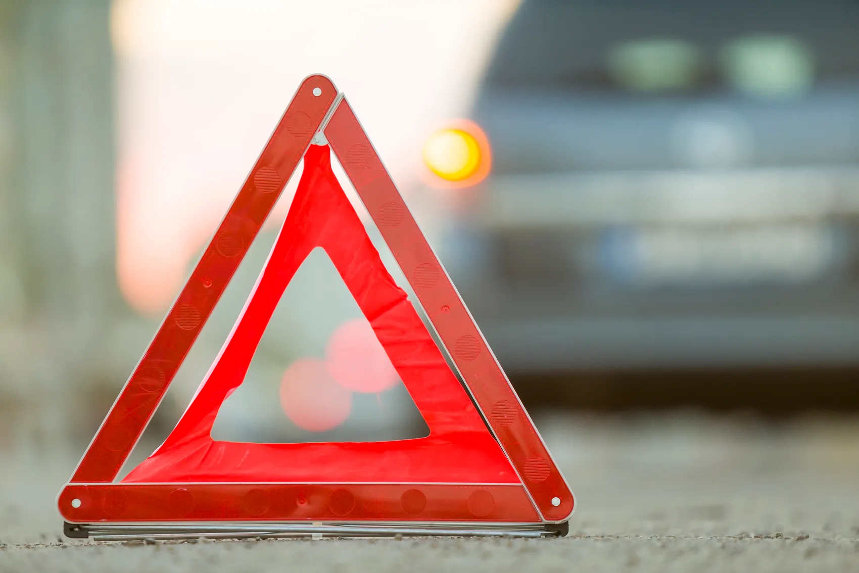 Rotes Warndreieck steht auf der Fahrbahn vor einem verschwommenen Auto, das vermutlich liegen geblieben ist.