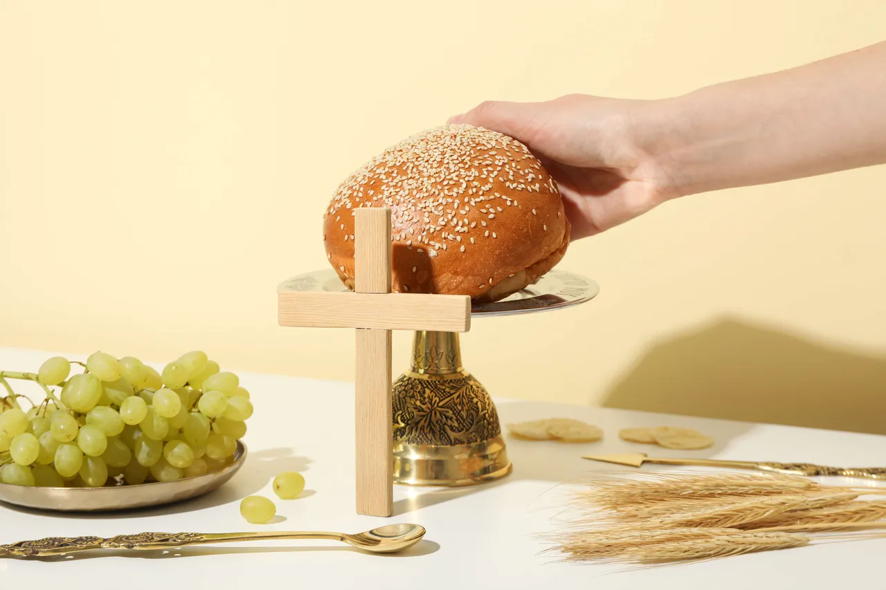 Ein Händchen legt ein mit Sesam bestreutes Brot auf ein kleines Holzkreuz über einem goldenen Kelch. Daneben liegen grüne Trauben, Hostien und Ähren.