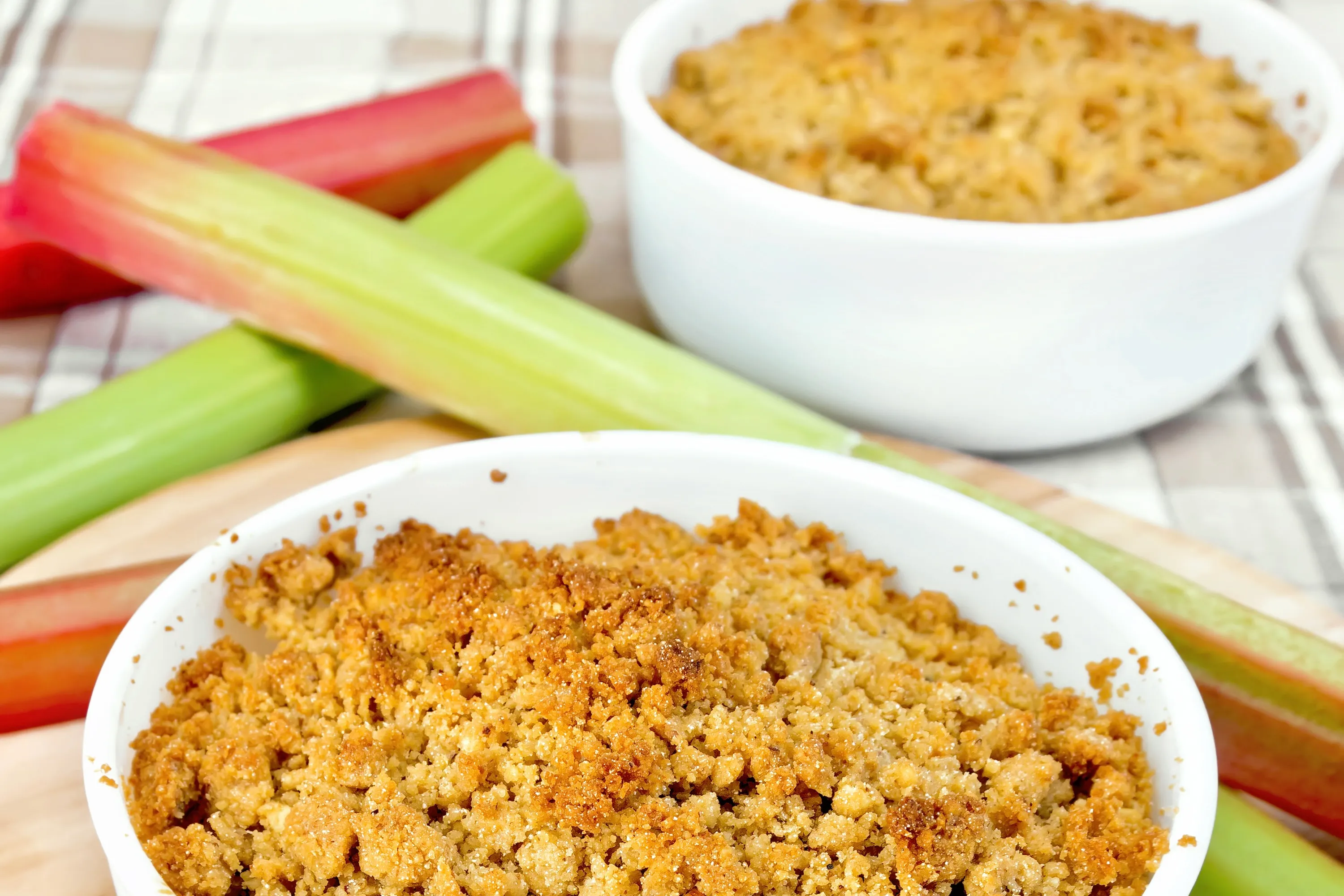 Zwei Schälchen Rhabarber-Crumble neben frischem Rhabarber auf einem Holzbrett.
