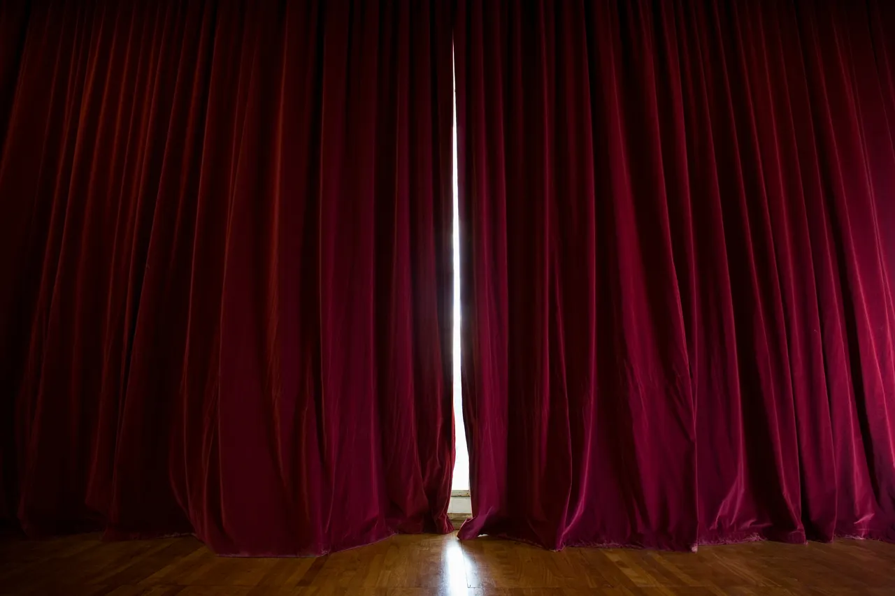 Ein geöffneter Vorhang aus rotem Samt gibt den Blick auf eine helle Bühne frei.