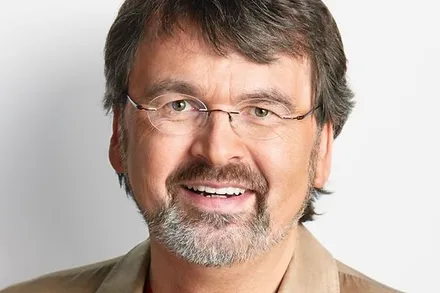 Ein Mann mit graumeliertem Haar und Bart lächelt in die Kamera. Er trägt eine Brille und ein hellbraunes Hemd.