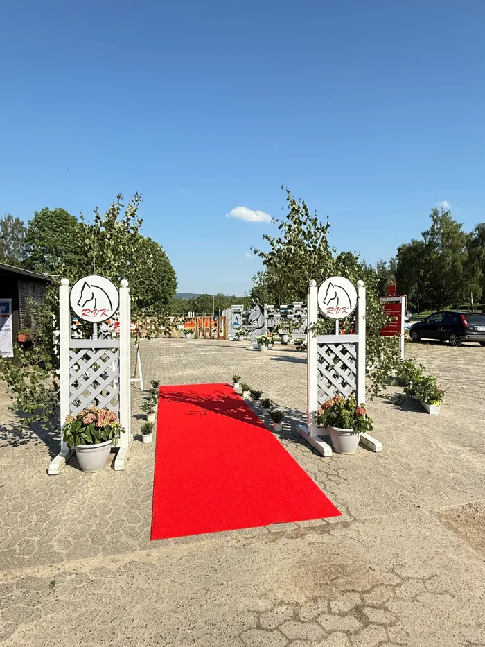 Ein roter Teppich führt zu einem Springparcours, flankiert von weißen Zäunen mit dem Logo des Reitvereins Kierspe. Im Hintergrund sind Gebäude, Bäume und Fahrzeuge zu sehen.