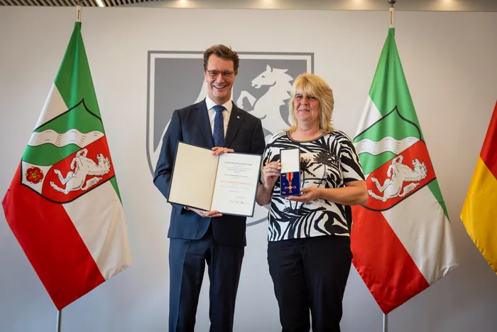 Hendrik Wüst überreicht Kathrin Tielmann-Lange eine Urkunde und das Bundesverdienstkreuz vor zwei Flaggen von Nordrhein-Westfalen.