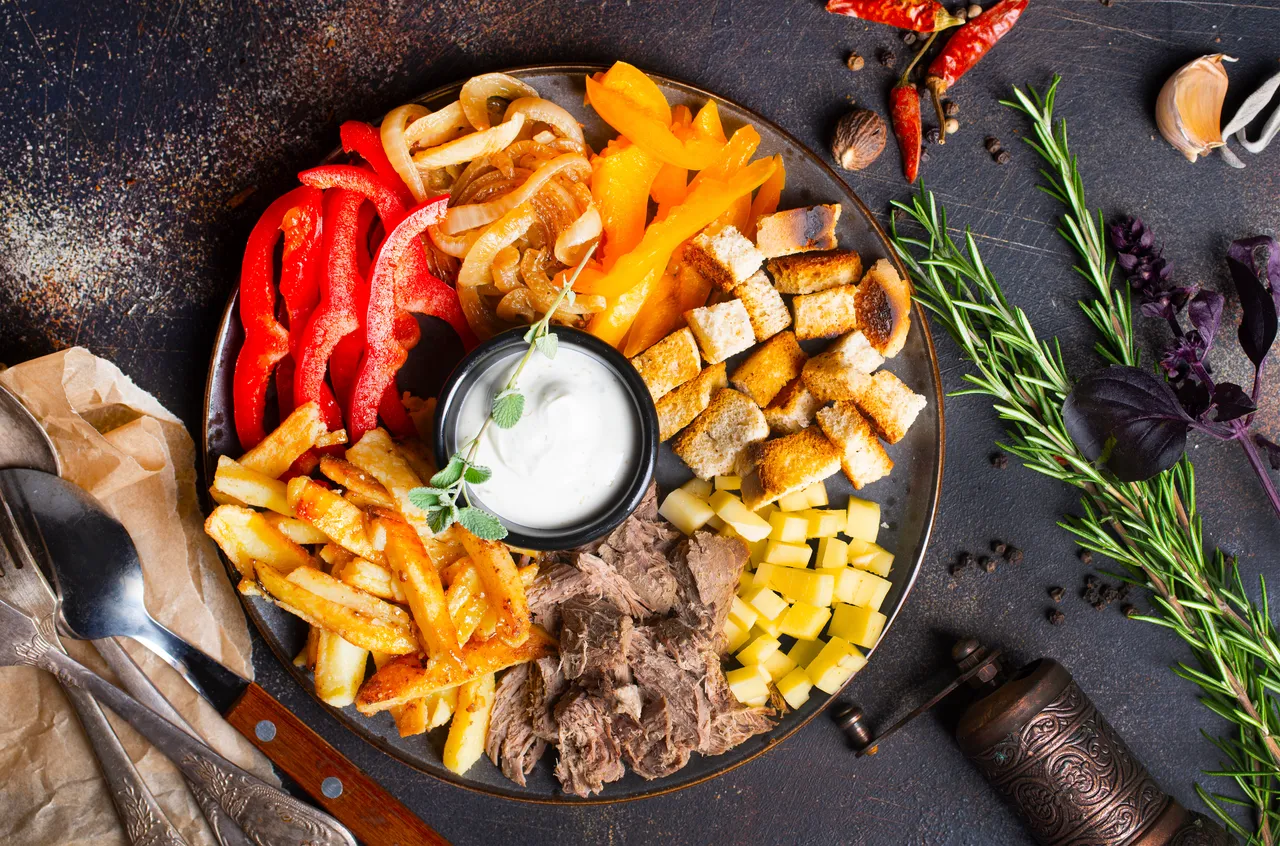 Verschiedene Beilagen wie Pommes, Paprika, Zwiebelringe, Brot und Fleisch auf einem Teller mit einer Schale Dip in der Mitte.