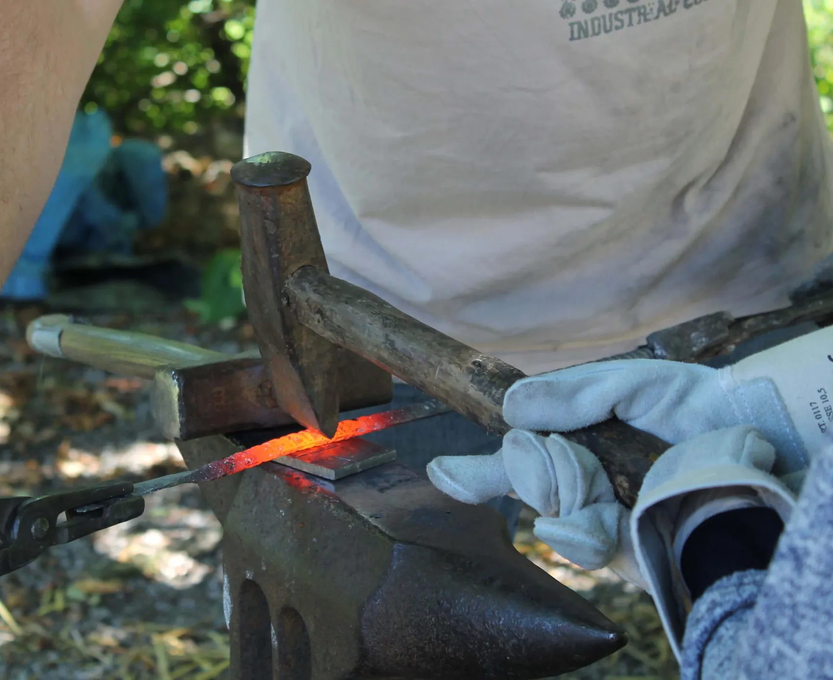 Ein Schmied bearbeitet mit Hammer und Zange ein glühendes Metallstück auf einem Amboss.