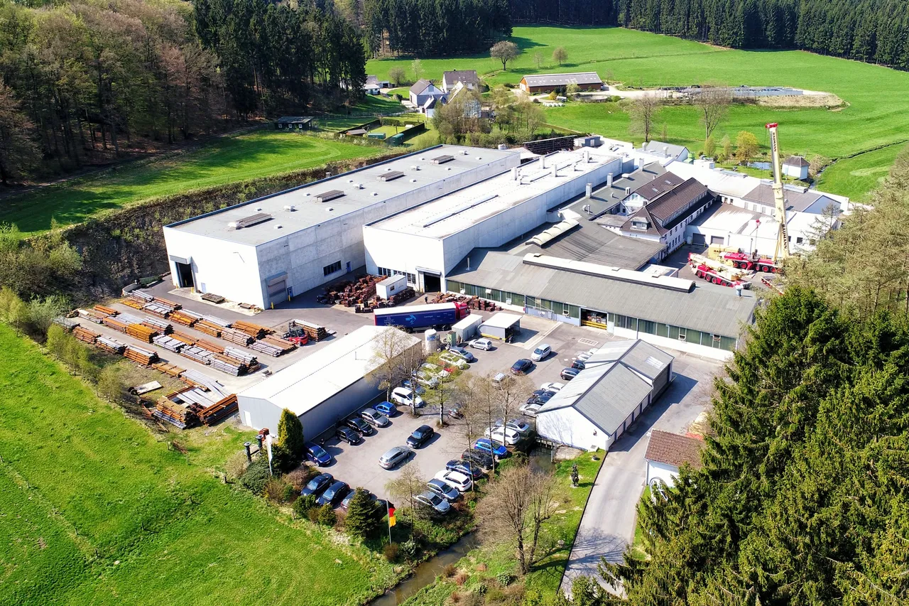 Ein Luftbild zeigt das Werksgelände der Schürfeld Umformtechnik GmbH mit mehreren Gebäuden, einem großen Parkplatz und Lagerflächen für Metallteile. Im Hintergrund sind Wiesen und ein Wald zu sehen.