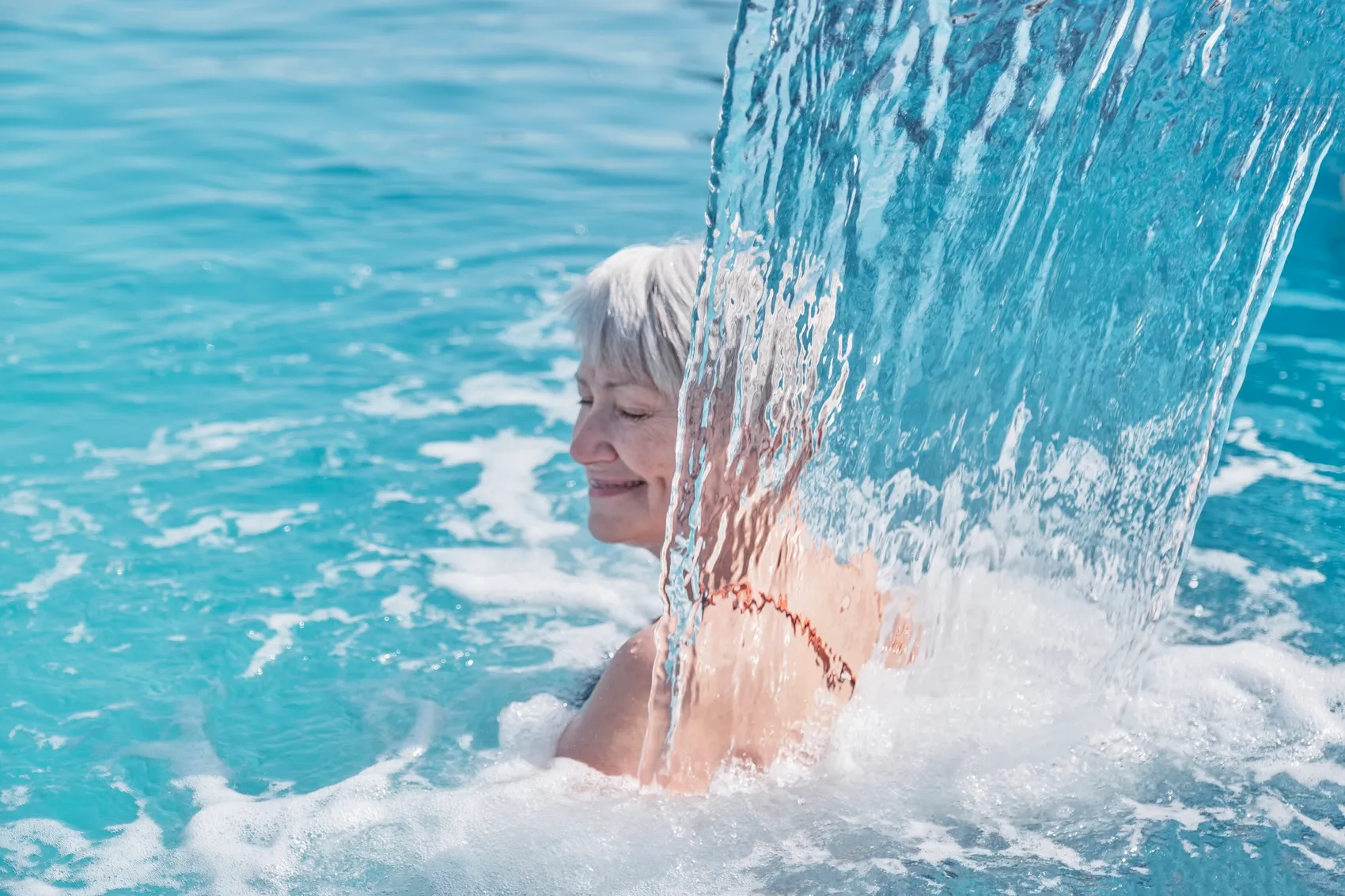 Ältere Frau lächelt entspannt, während sie unter einem Wasserfall in einem Pool steht.