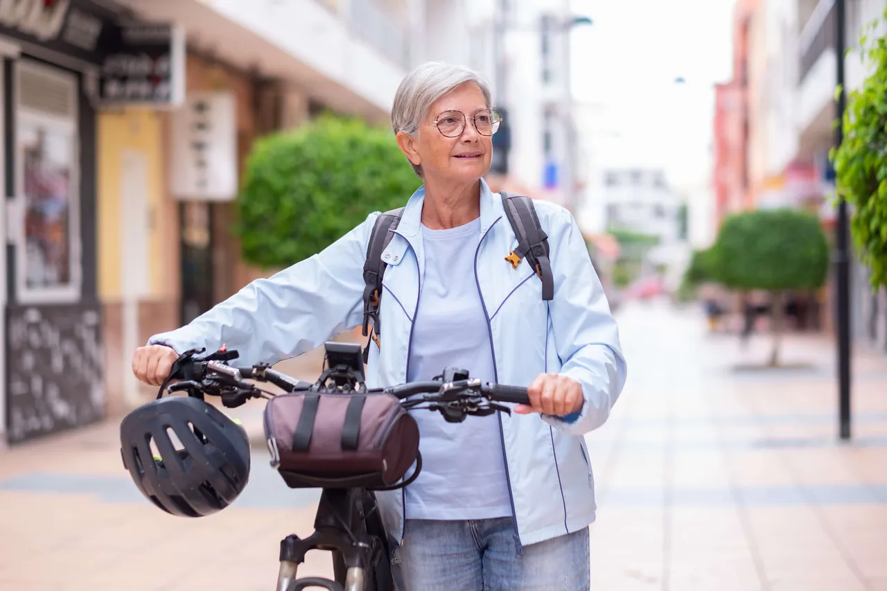 Eine Seniorin mit Brille und hellblauer Jacke steht mit einem Fahrrad und einem am Lenker hängenden Helm in einer sonnigen Fußgängerzone.