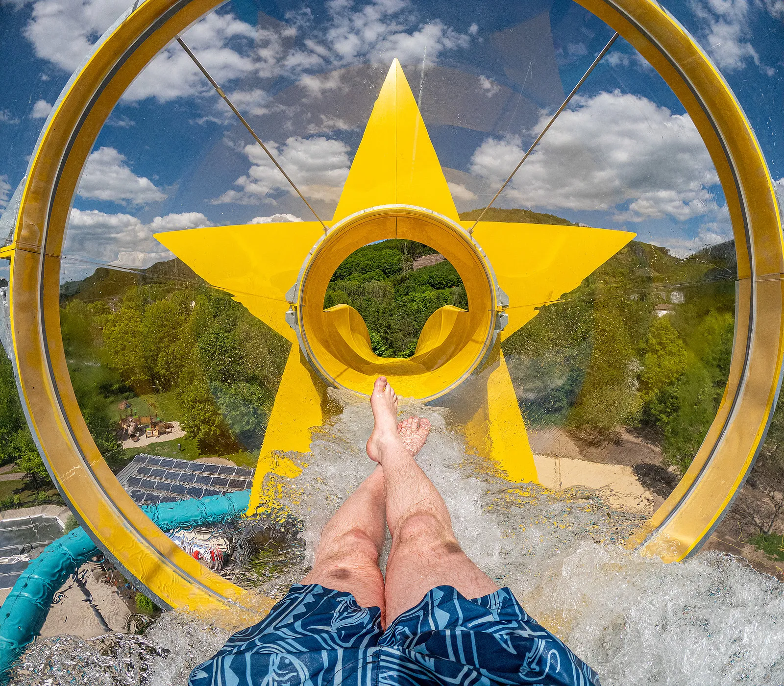 Person rutscht auf einer gelben Wasserrutsche in Sternform, umgeben von transparentem Tunnel und blauem Himmel.