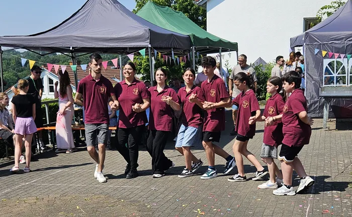 Eine Gruppe von Menschen in roten Poloshirts mit gelbem Logo tanzt im Freien vor mehreren Pavillons. Im Hintergrund sind weitere Besucher und Girlanden zu sehen.
