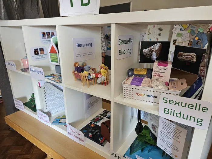 Ein weißes Regal ist mit Ordnern und Informationsmaterialien zu Themen wie Beratung, sexueller Bildung, Traumapädagogik und Hygieneboxen gefüllt. Im Vordergrund steht eine kleine Puppenhaus-Szene mit einer Frau und einem Kind.