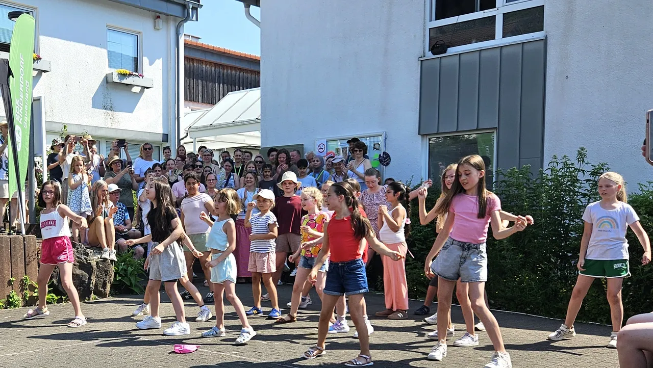 Eine Gruppe von Kindern und Jugendlichen tanzt im Freien vor einem Gebäude, während im Hintergrund Zuschauer stehen und zusehen.
