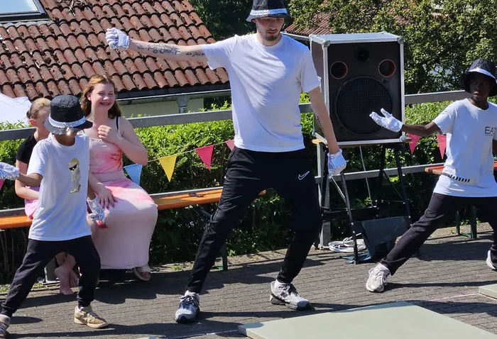 Eine Frau in einem rosa Kleid und ein Mann in Sportkleidung tanzen auf einer Bühne vor Kindern mit silbernen Accessoires. Im Hintergrund sind Zuschauer und festliche Girlanden zu sehen.