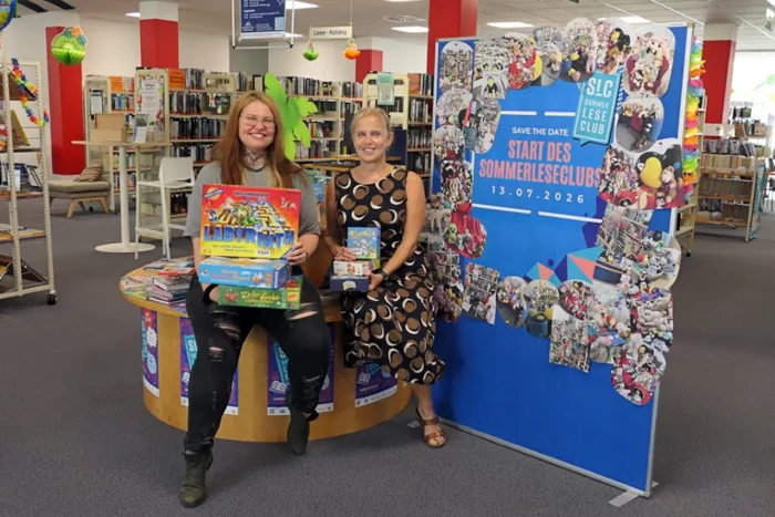 Katharina Fuhrmann und Christiane Bauckhage sitzen in einer Stadtbücherei und halten Brettspiele vor einem blauen Banner für den Sommerleseclub.