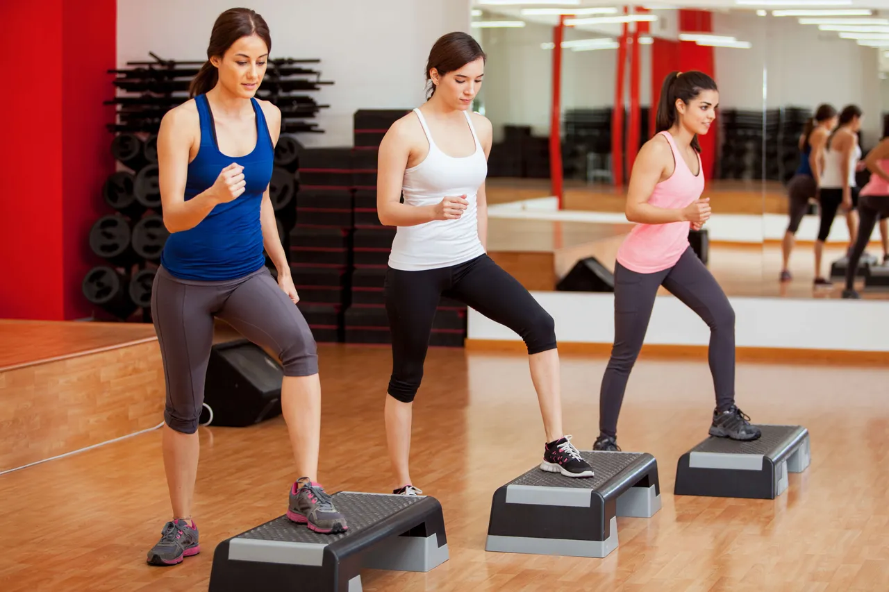 Drei Frauen in Sportkleidung führen Übungen auf Step-Plattformen in einem Fitnessstudio aus.