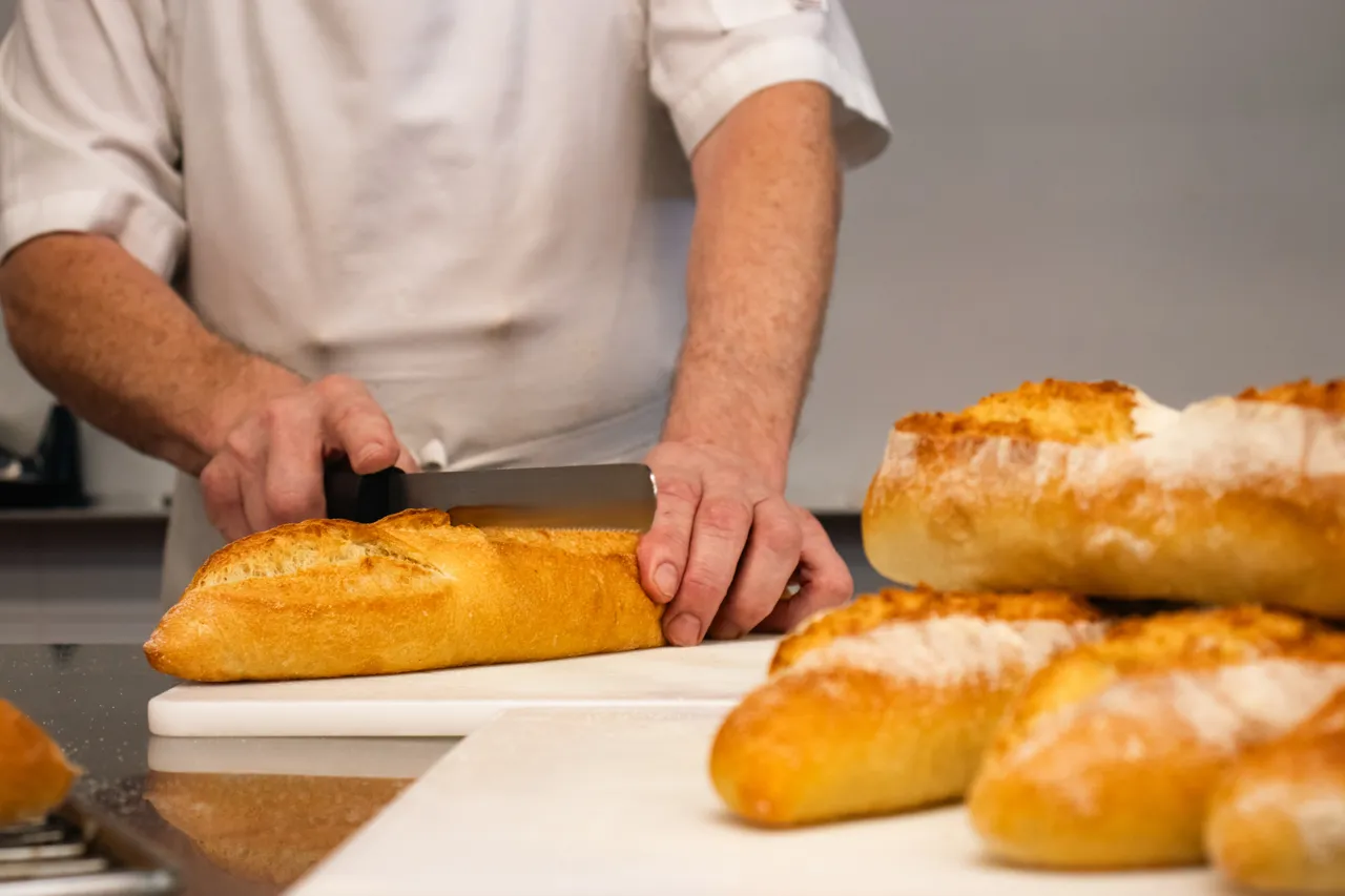 Ein Koch schneidet mit einem Messer ein Baguette auf einem weißen Schneidebrett. Im Hintergrund liegen weitere Brote bereit.