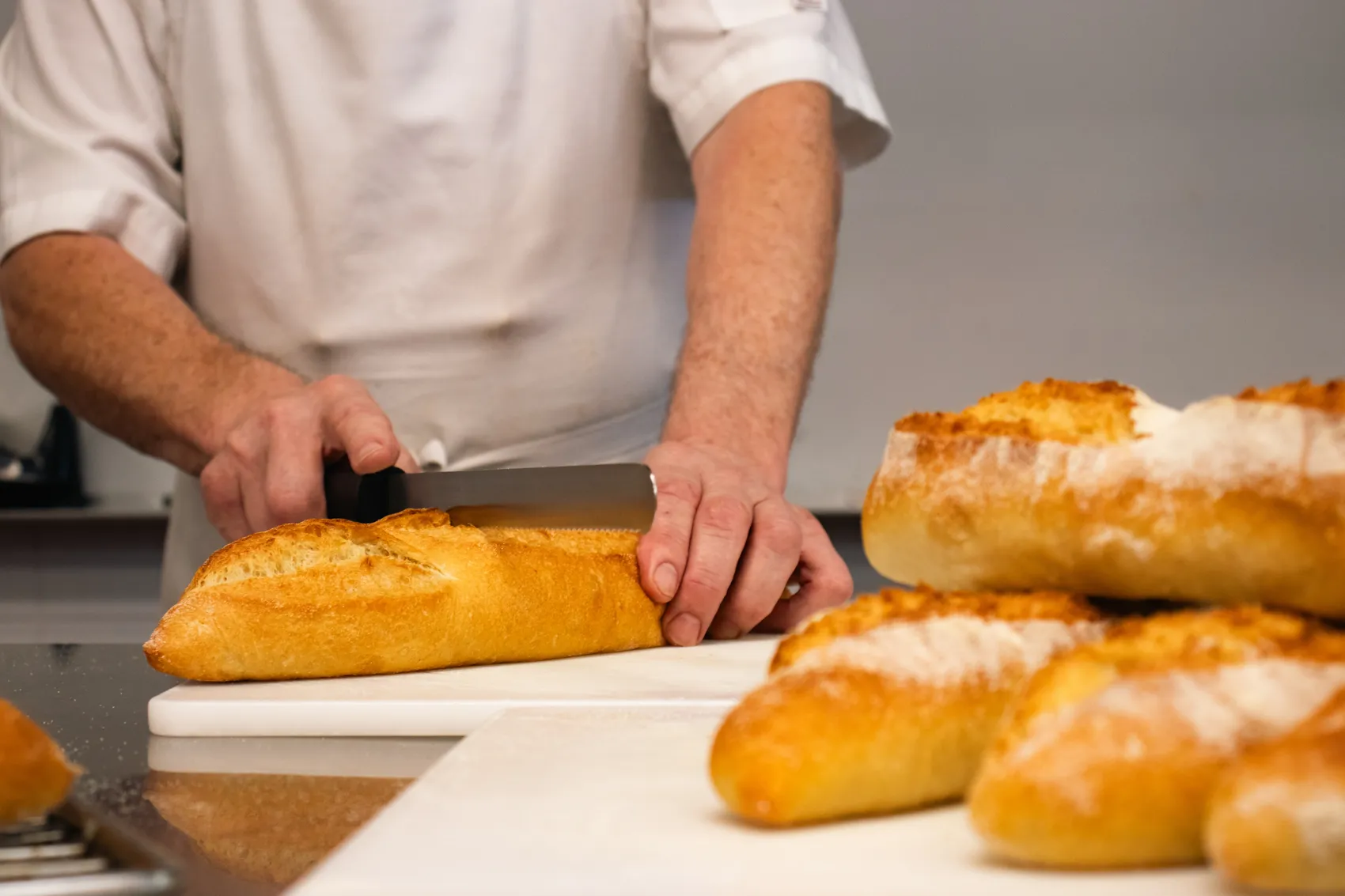 Ein Koch schneidet mit einem Messer ein Baguette auf einem weißen Schneidebrett. Im Hintergrund liegen weitere Brote bereit.