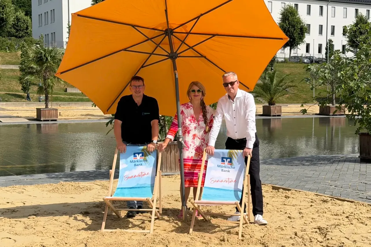 Thomas Bielawski, Silke Weidenheimer und Artur Merz stehen unter einem großen orangefarbenen Sonnenschirm auf einer Sandfläche am Wasser.