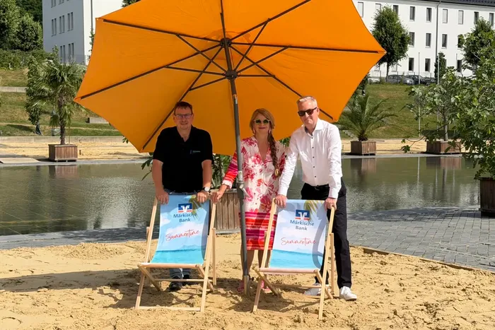 Thomas Bielawski, Silke Weidenheimer und Artur Merz stehen unter einem großen orangefarbenen Sonnenschirm auf einer Sandfläche am Wasser.