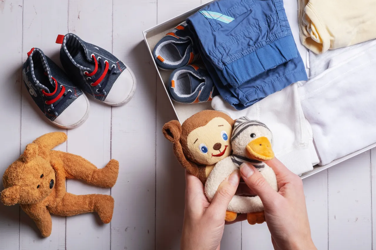 Blaue Babyschuhe, ein Teddybär und eine Kiste mit Babykleidung liegen auf einem Holztisch, während Hände zwei kleine Stofftiere halten.