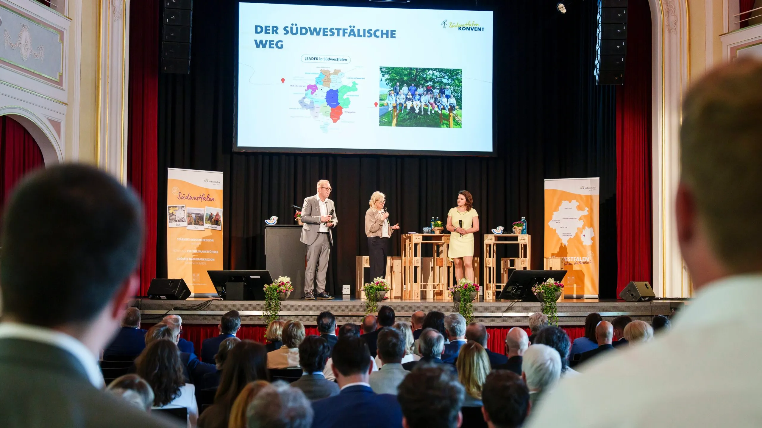 Drei Personen stehen auf einer Bühne vor einer Präsentation mit der Aufschrift „Der Südwestfälische Weg“ und einer Karte der Region Südwestfalen. Im Publikum sitzen zahlreiche Zuhörer.