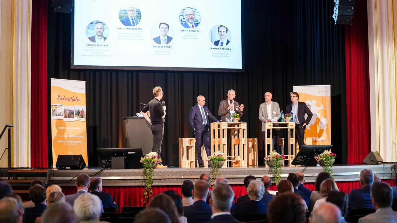 Auf einer Bühne diskutieren fünf Männer vor einem Publikum. Im Hintergrund werden Porträts und Namen von Personen auf einer Leinwand angezeigt.