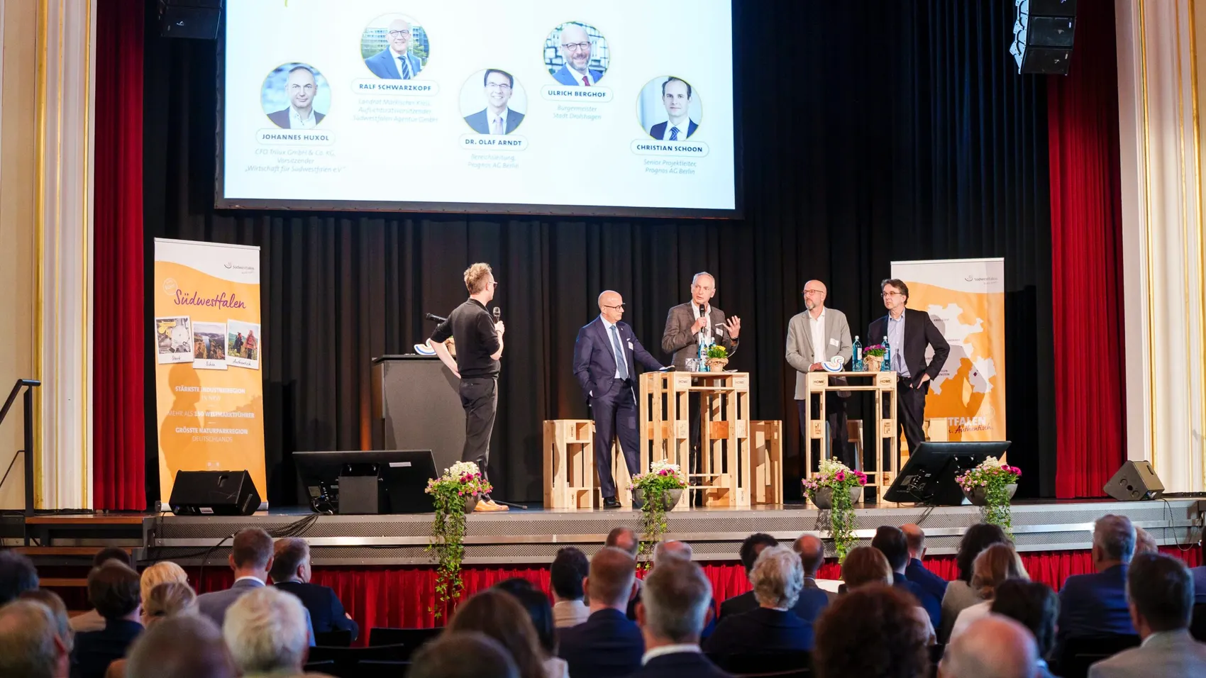 Auf einer Bühne diskutieren fünf Männer vor einem Publikum. Im Hintergrund werden Porträts und Namen von Personen auf einer Leinwand angezeigt.