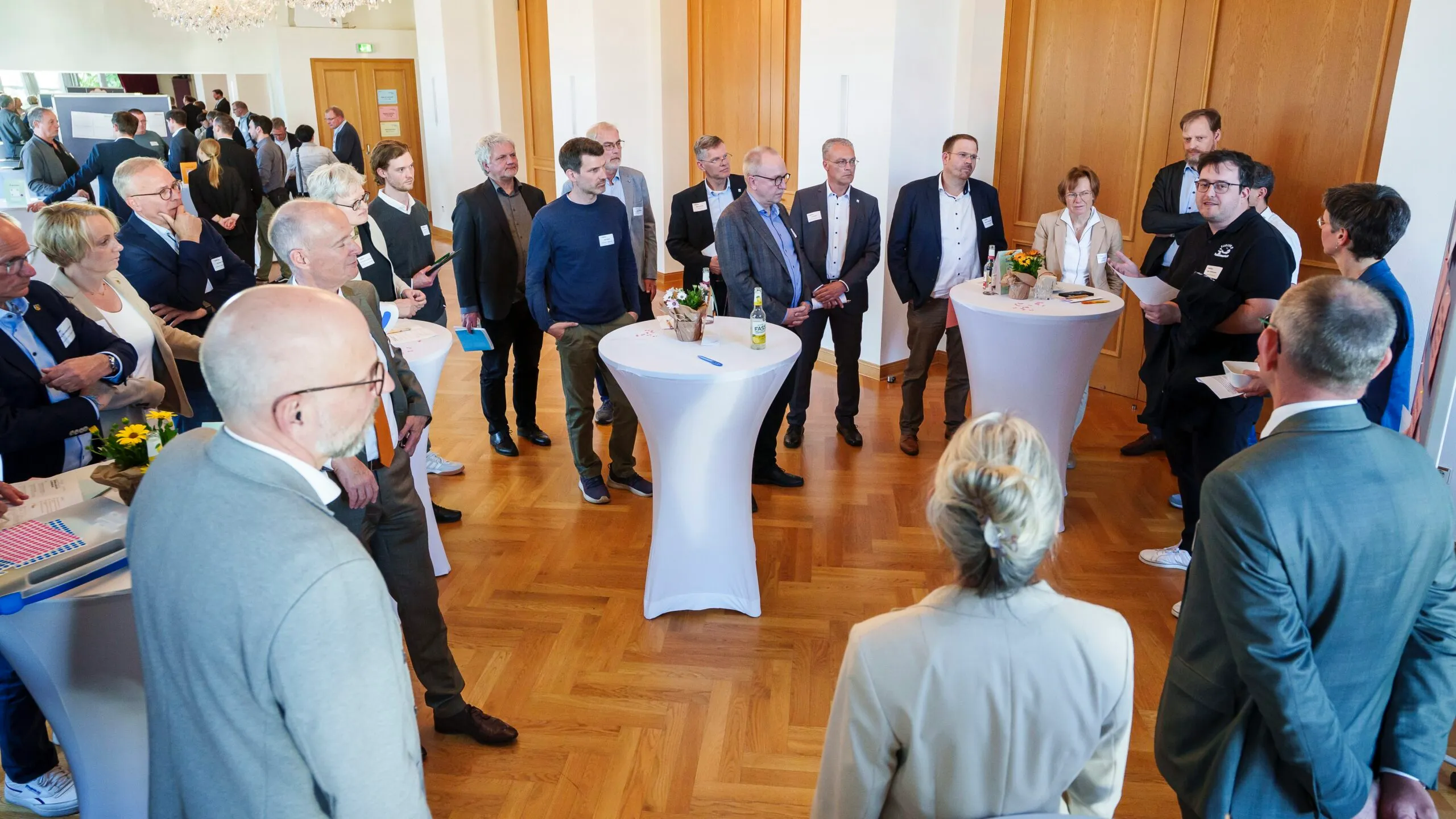 Eine Gruppe von etwa dreißig Personen steht und unterhält sich in einem hellen Raum mit Parkettboden. Einige stehen um Stehtische, andere bilden kleine Gesprächsrunden.