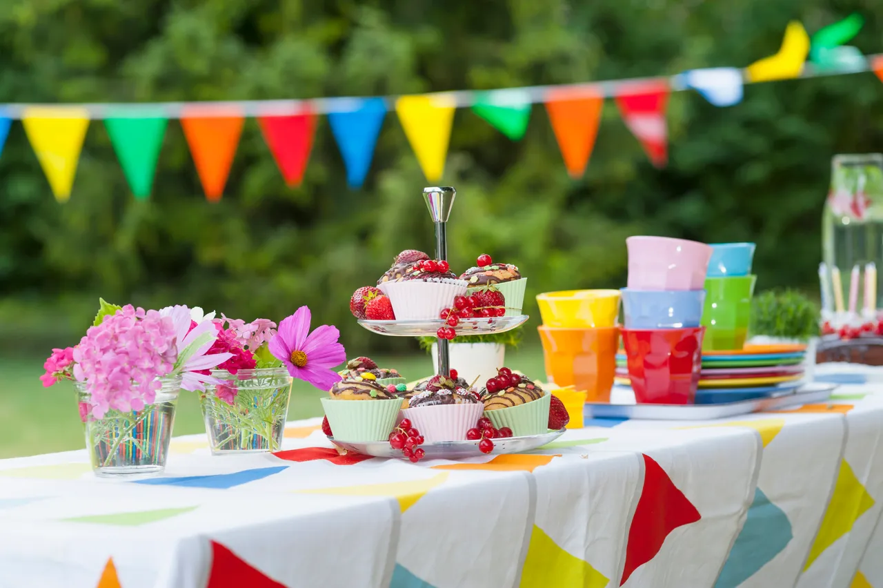 Festlich gedeckter Tisch im Freien mit Cupcakes, Beeren, bunten Bechern und Blumenarrangements für eine Sommerparty.