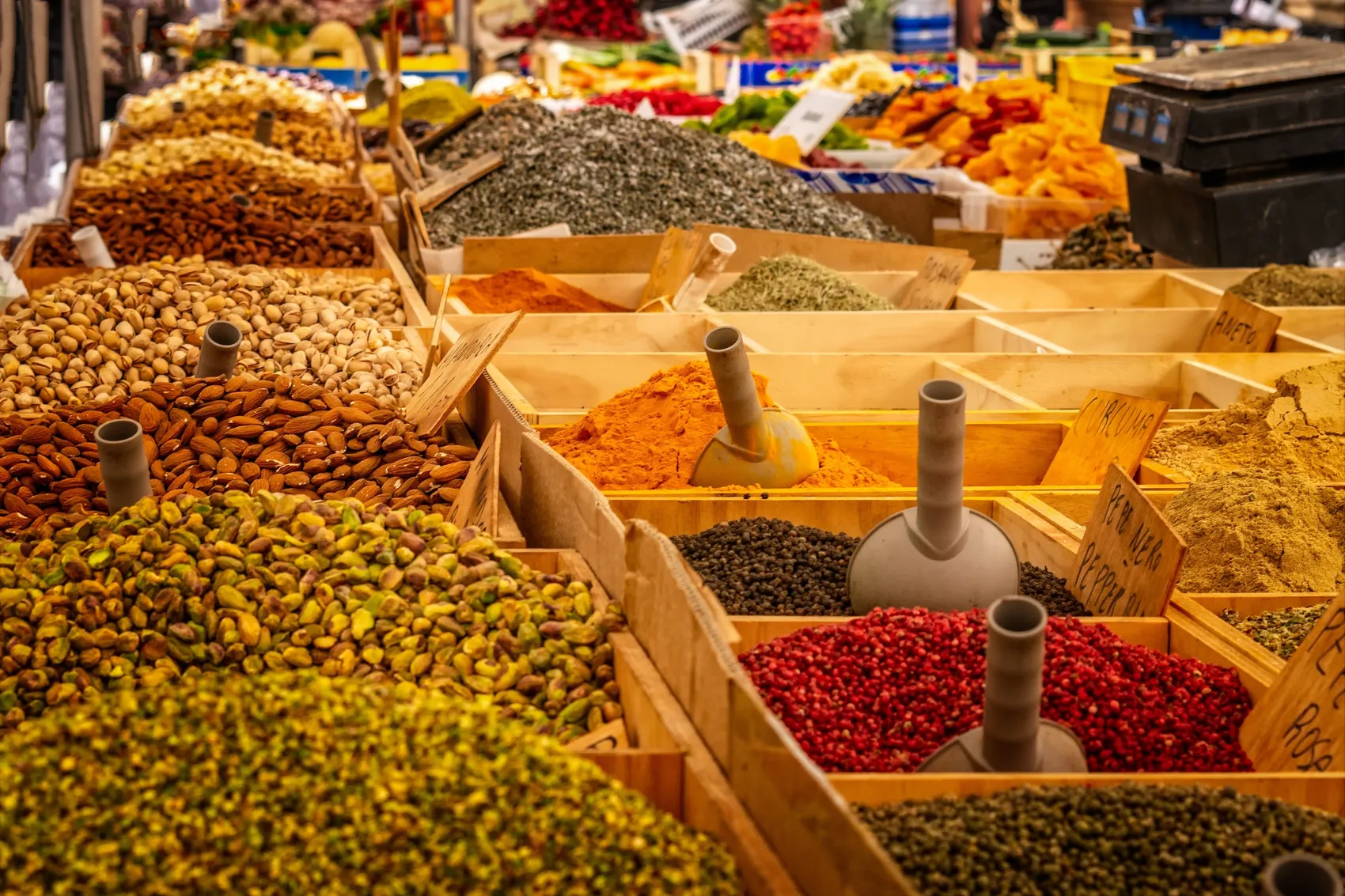Verschiedene Schüttungen mit Nüssen, Gewürzen und Kräutern stehen auf einem Marktstand bereit. Im Vordergrund sind Mandeln, Pistazien, gelbes Currypulver und rote Paprikaflocken zu sehen.