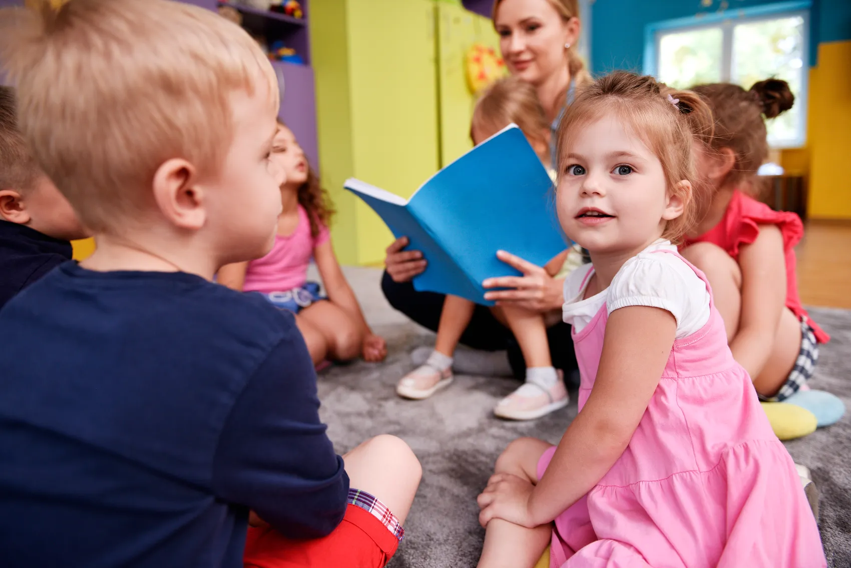 Eine Lehrerin liest einer Gruppe von Kindern in einem bunten Raum aus einem blauen Buch vor.