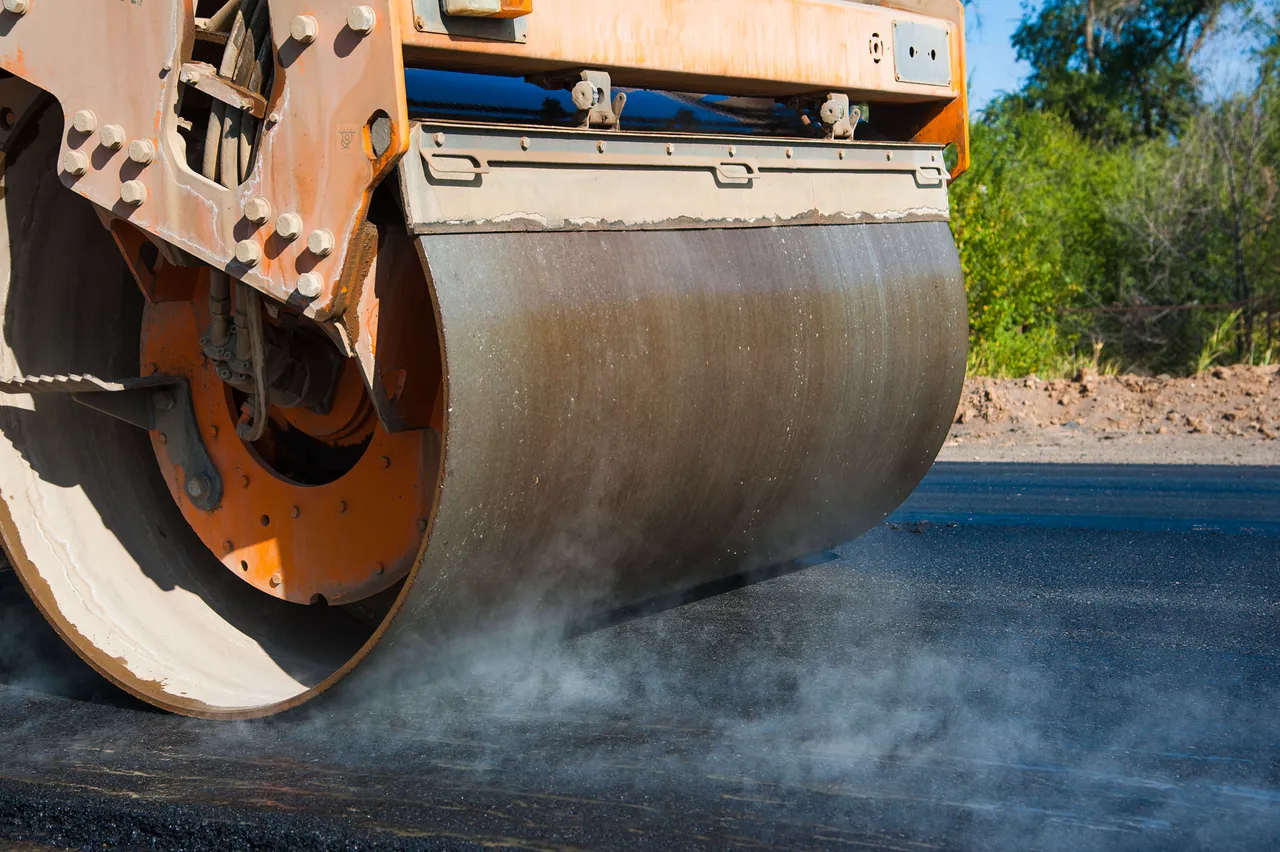 Eine orangefarbene Straßenwalze verdichtet Asphalt auf einer neuen Fahrbahn. Dampf steigt auf.
