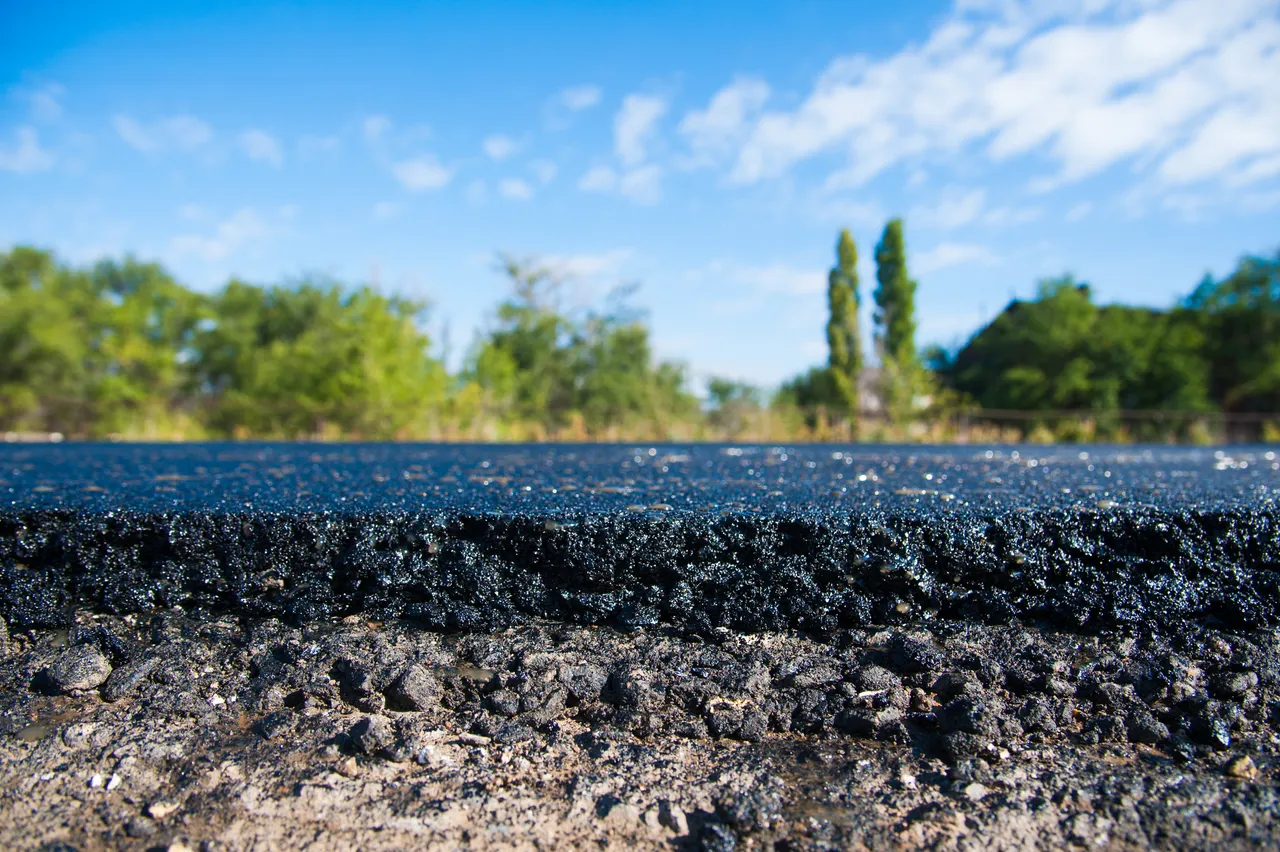 Nahaufnahme der rauen Textur einer frisch asphaltierten Straße vor einem Hintergrund aus Bäumen und blauem Himmel.