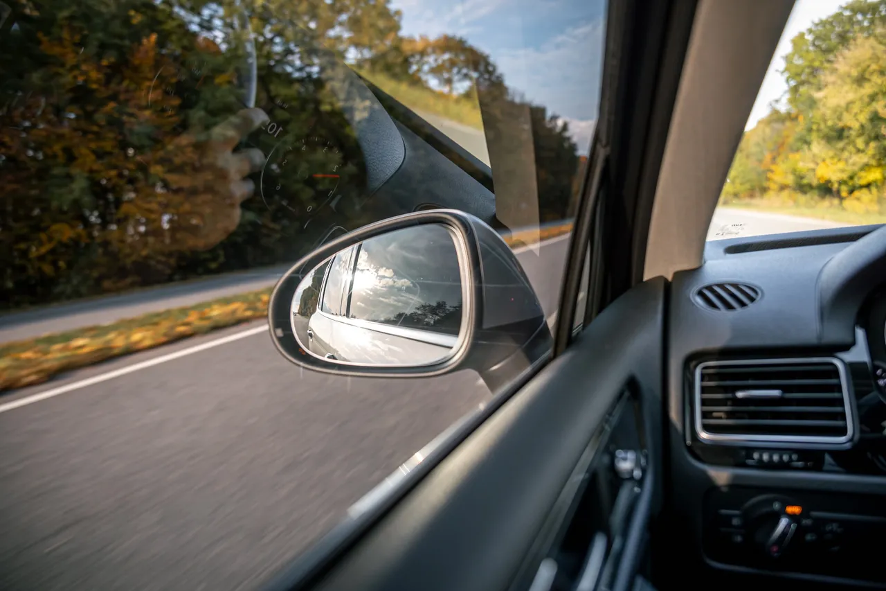 Blick aus dem Auto auf den Außenspiegel, der die Straße und den Himmel hinter dem Fahrzeug reflektiert.