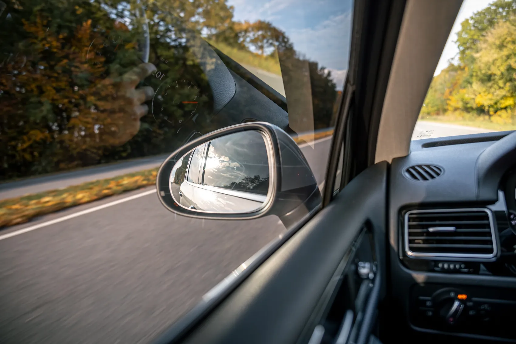 Blick aus dem Auto auf den Außenspiegel, der die Straße und den Himmel hinter dem Fahrzeug reflektiert.